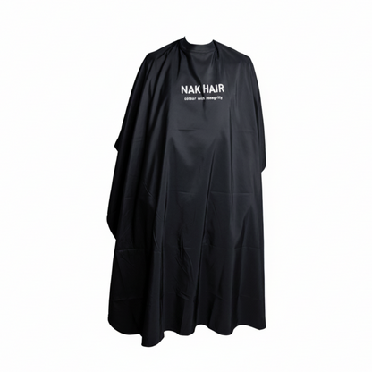 A black salon cape with 'NAK HAIR' printed in white on the chest, displayed against a solid pale pink background.